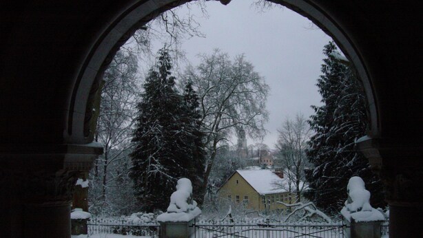 Paisaje nevado visto a través de una ventana arqueada, con árboles cargados de nieve y un edificio con tejado cubierto de nieve.