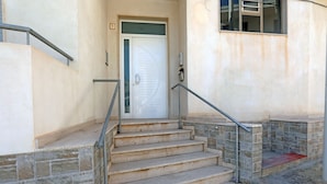 A set of concrete steps leading to a white door with a glass panel, a metal handrail, and a stone wall on one side. - Appartamento Calipso (Santa Croce Camerina)