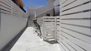 Un estrecho pasillo exterior con una cerca de metal blanco, una bicicleta y una escalera que conduce a un edificio.