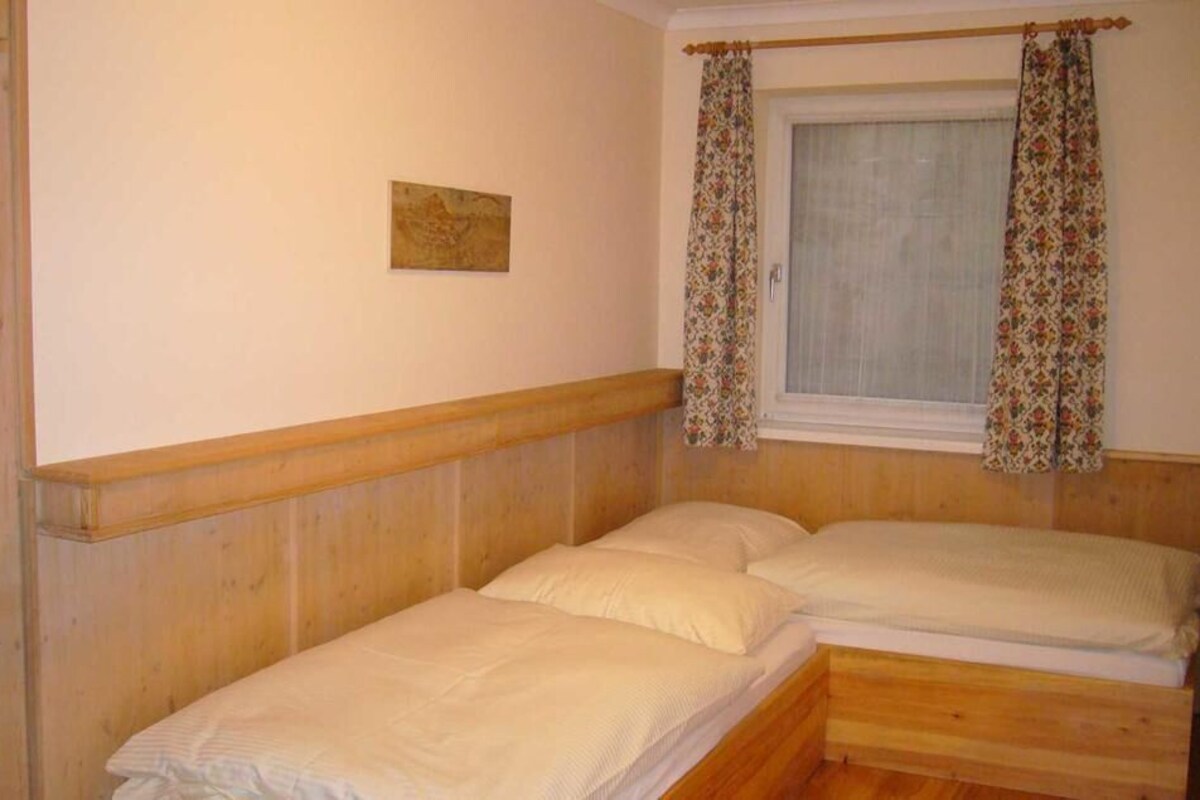 Una habitación pequeña con cama individual, cabecera de madera, ventana con cortinas florales y un cuadro pequeño en la pared.