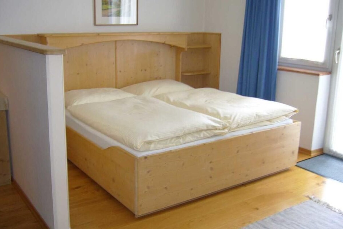 Una cama individual con cabecera de madera y colchón blanco en habitación con suelo de madera y cortina azul.