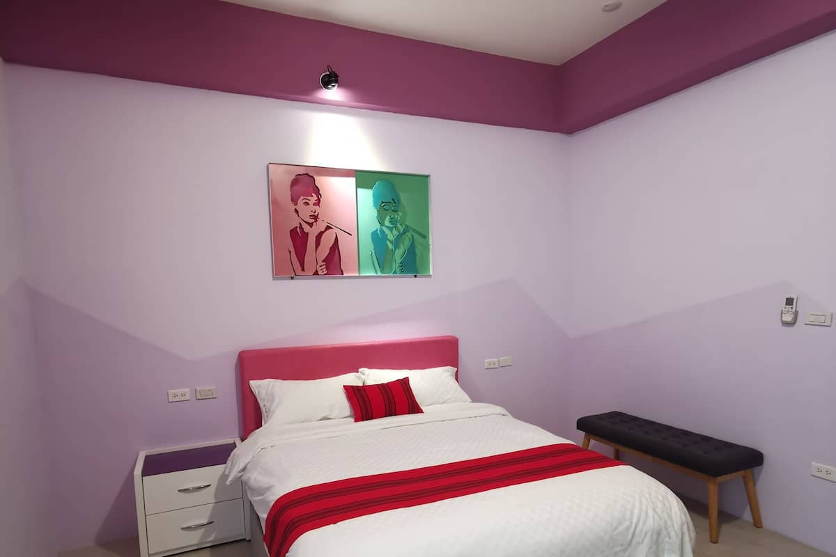 A bedroom with a bed, a nightstand, a bench, and two wall-mounted light switches.