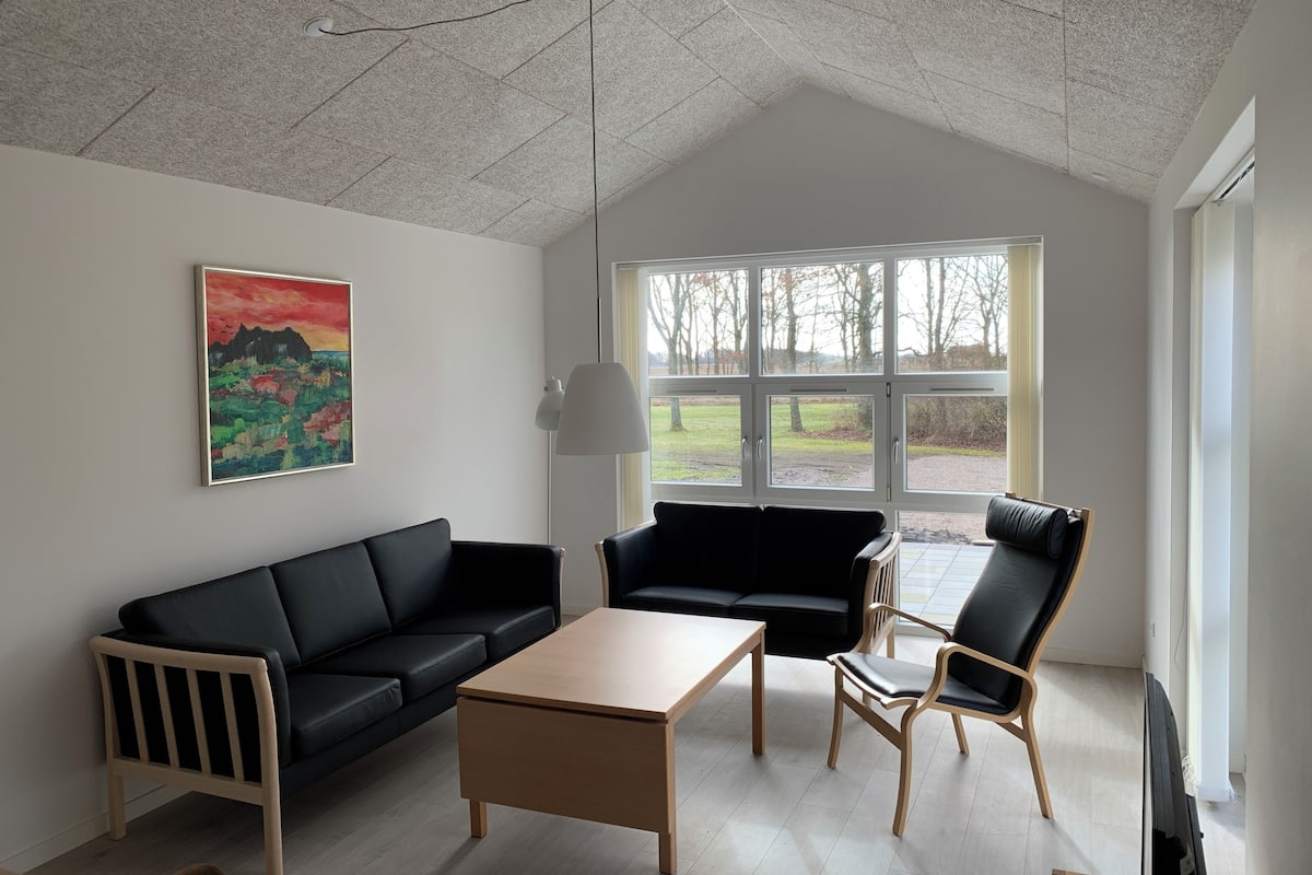 A modern living room with a black sofa, a wooden coffee table, and a black armchair. There is a painting on the wall and a window with a view of the outdoors.