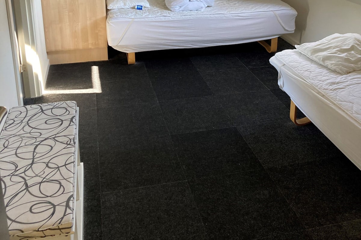 A small, dimly lit room with a sloped ceiling, a wooden wardrobe, two beds, and a patterned rug.