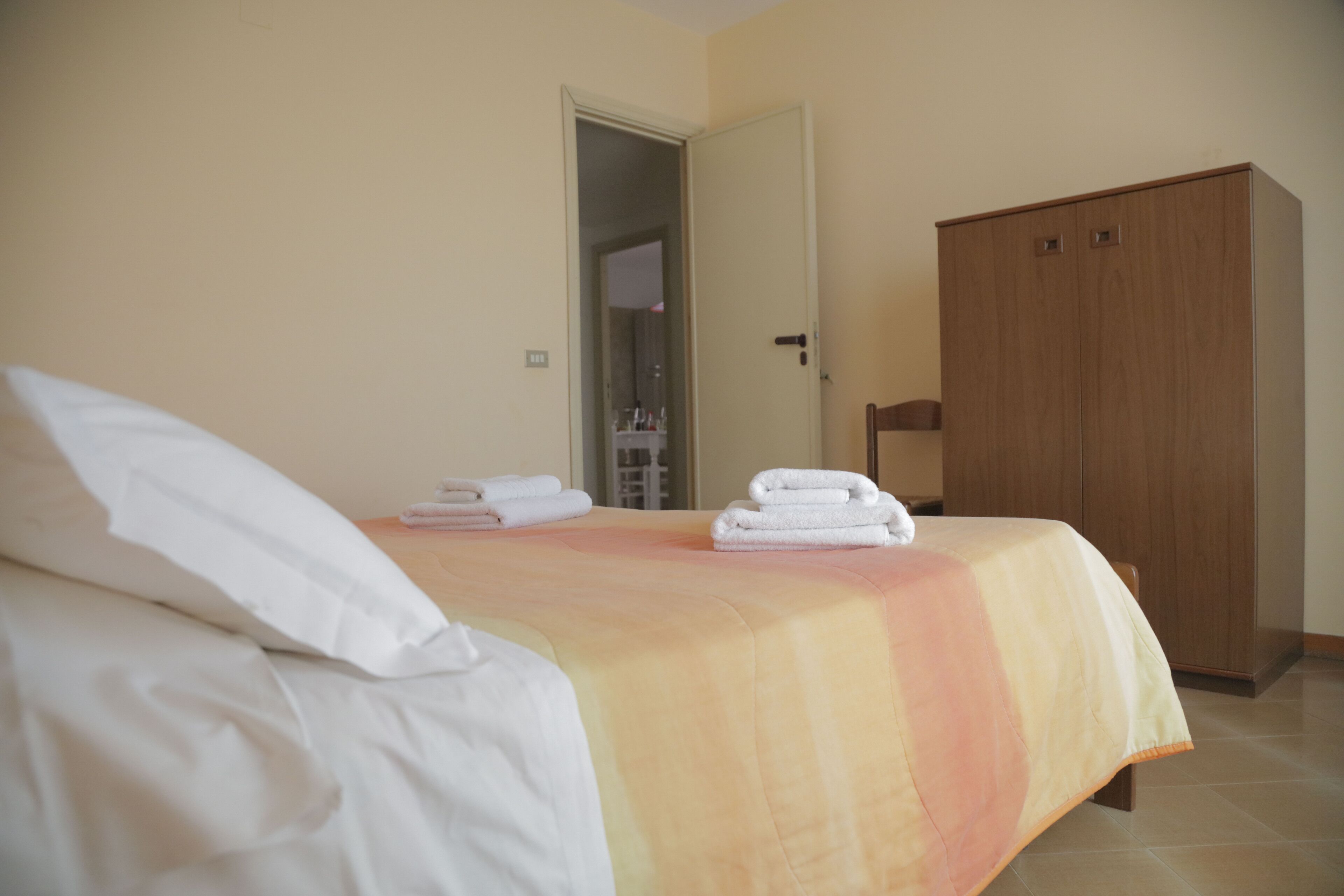 Un lit bien fait, avec du linge de lit blanc et une couvre-lit de couleur pêche, une armoire en bois et une porte donnant sur une salle de bain.