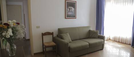 A living room with a sofa, a chair, a vase with flowers, a framed picture on the wall, and a door leading to another room.