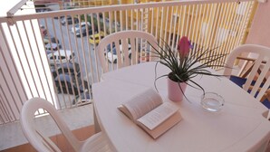 Un balcón con una mesa blanca, sillas, un libro, una planta en maceta y un vaso de vidrio transparente.
