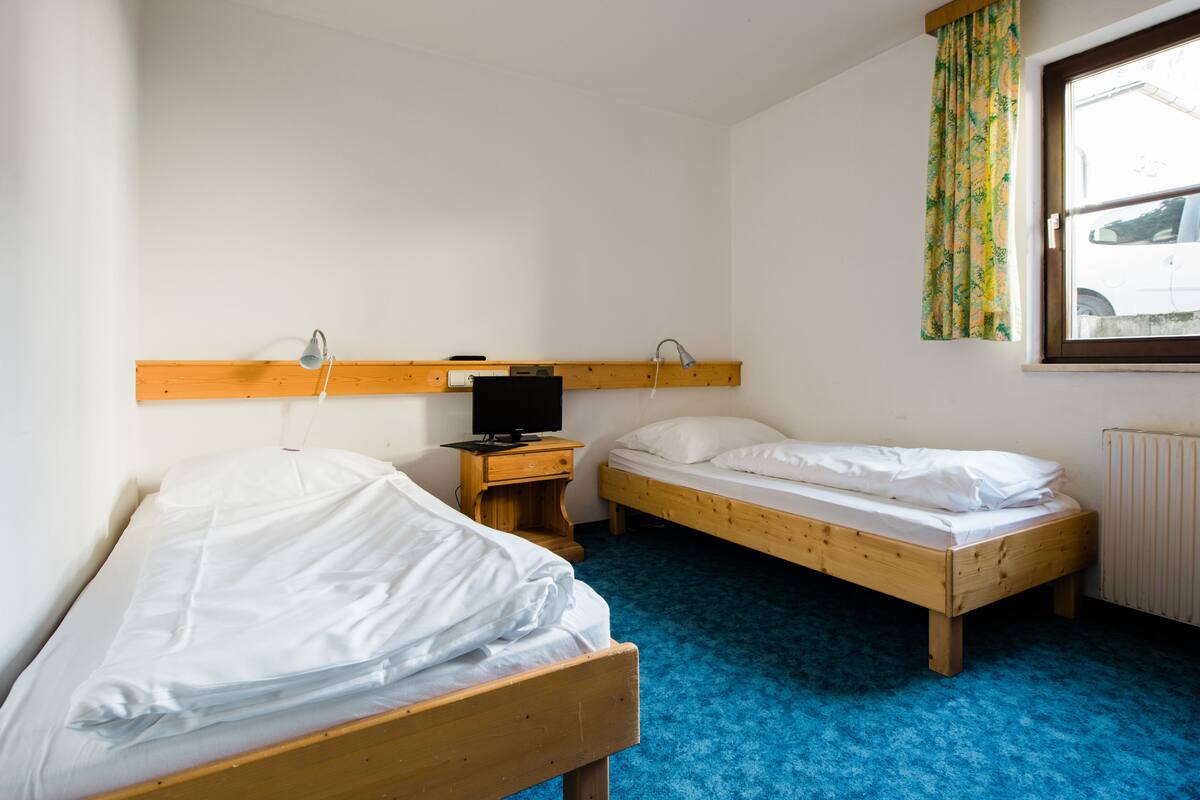 A room with two wooden beds, a small wooden nightstand with a television, and a window with a curtain.