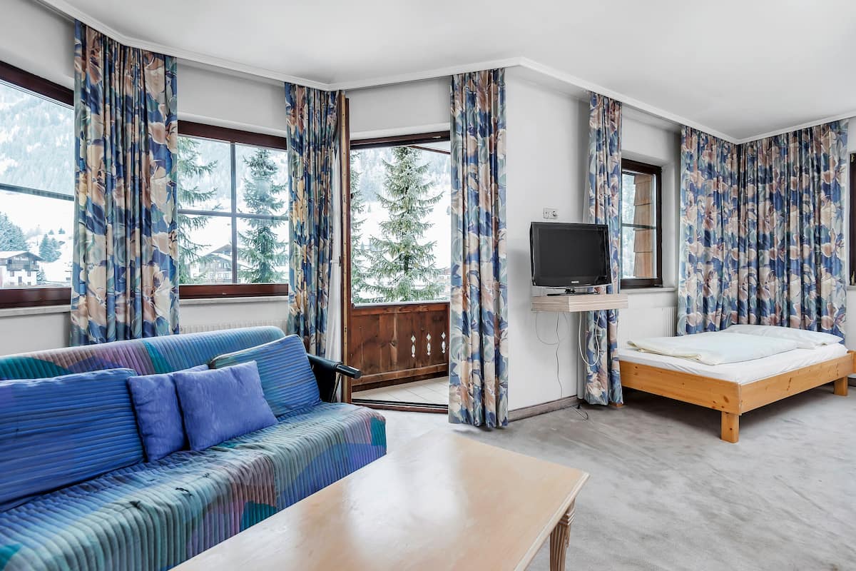 A room with a sofa, a bed, a TV, and large windows with curtains.
