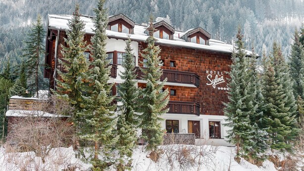 Un hôtel à plusieurs étages, doté d’une façade en bois, de balcons et offrant une vue sur des arbres et des montagnes enneigés.