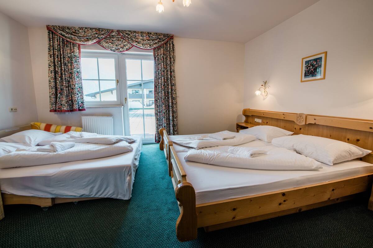 Camera d'albergo con due letti singoli, una testiera in legno, una finestra con tende floreali, un termosifone e un piccolo fiorileto a parete.