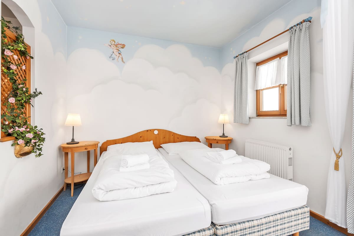 Cama doble con sábanas blancas, dos mesitas de noche con lámparas, una ventana con cortinas y una pared con un mural de nubes.