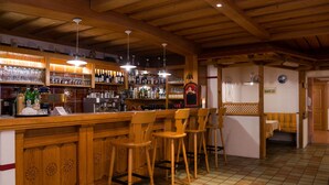 Uma área de bar com balcão de madeira, várias prateleiras com garrafas e copos, e três banquetas de madeira.