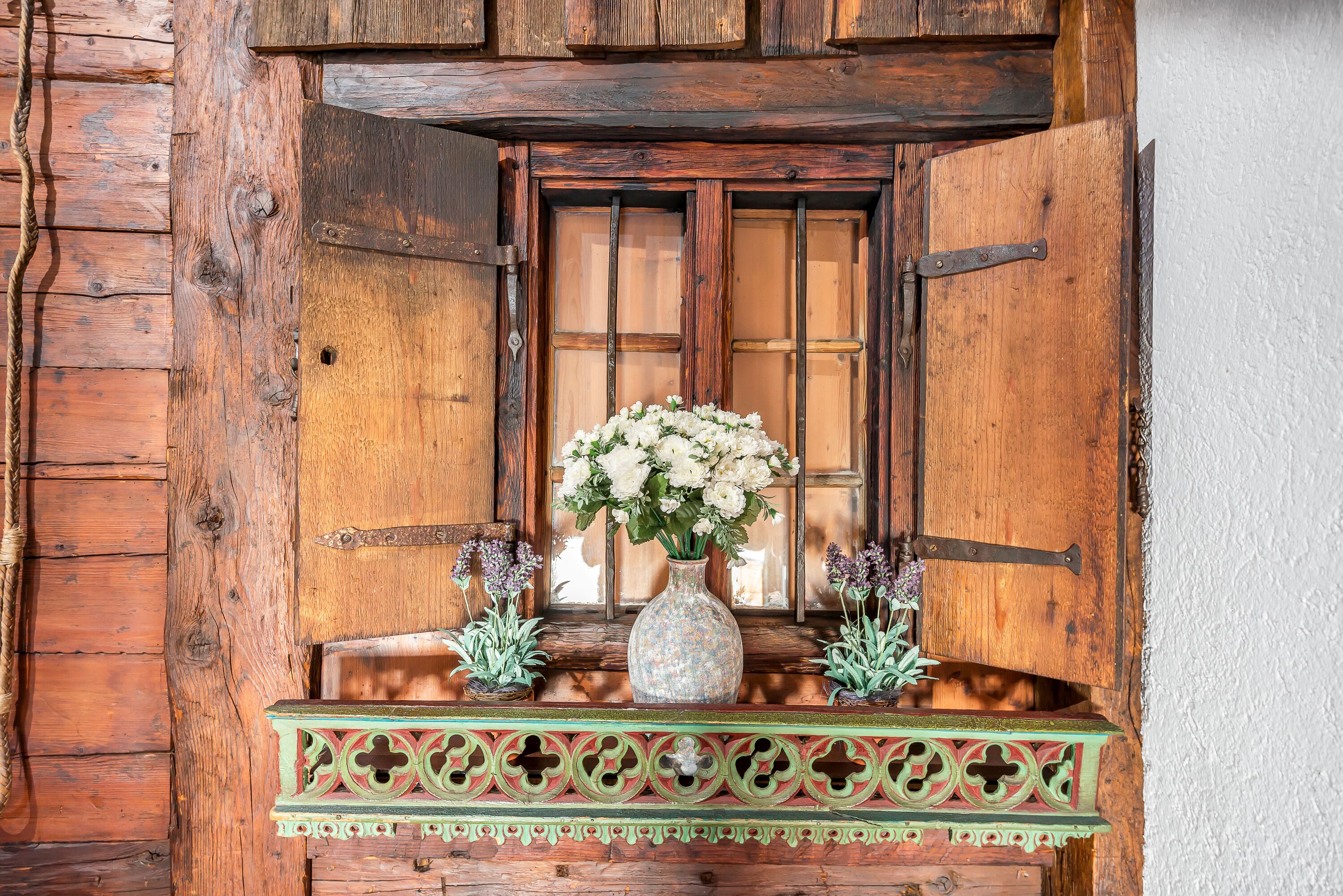 Une fenêtre en bois rustique, ornée d’un vase de fleurs blanches et de deux petites plantes en pot, le tout posé sur une étagère décorative.