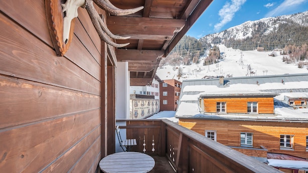 A wooden balcony with a table and chair, overlooking a snowy mountain and buildings. - Heart Hotel Talhoff Nigg (Sankt Anton am Arlberg)