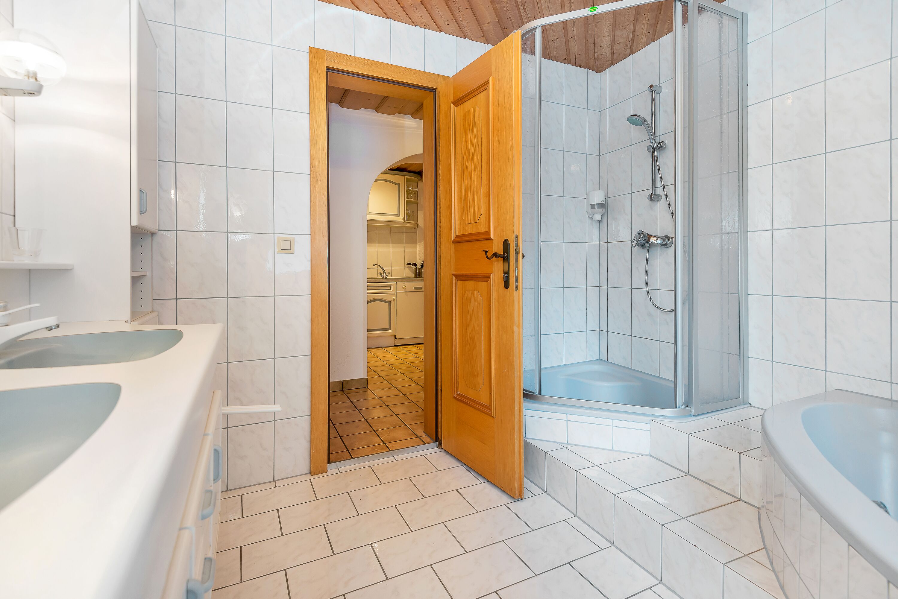 A modern bathroom with a shower, two sinks, and a wooden door leading to a kitchen.