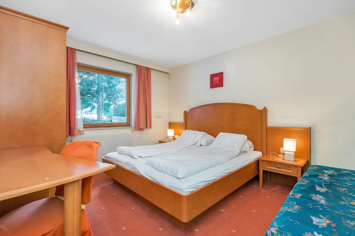 Une chambre d’hôtel avec un lit, un bureau, une chaise, une fenêtre avec des rideaux, une table de chevet avec une lampe et un panneau rouge sur le mur.
