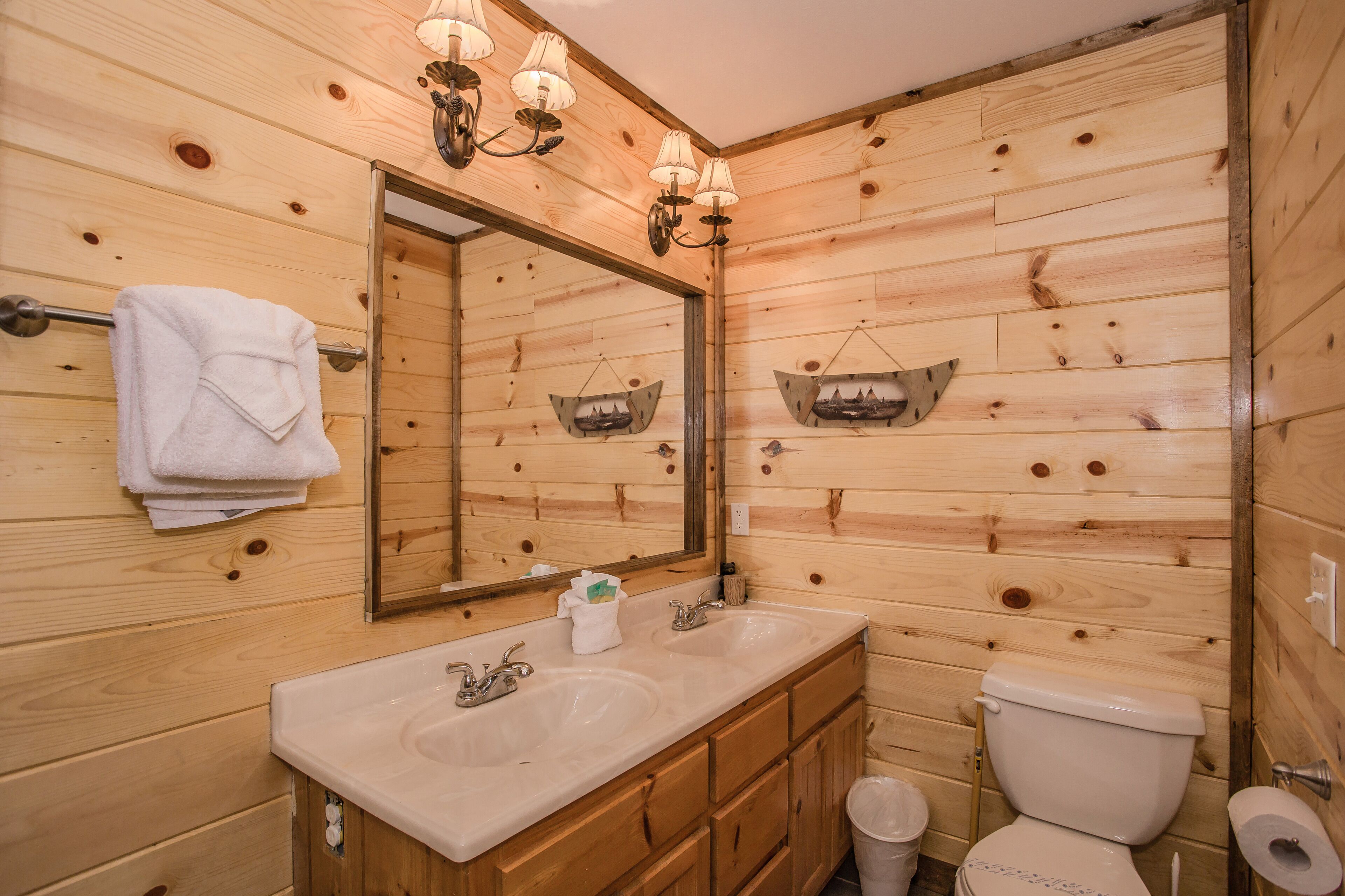 A wooden cabin-style bathroom with a double sink, a toilet, and a mirror with wall-mounted lights.
