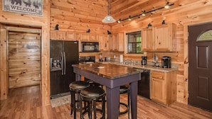 Ein Interieur einer Holzhütte mit einer Küche, die eine zentrale Kochinsel, Barhocker, einen Kühlschrank, eine Mikrowelle und eine Kaffeemaschine beinhaltet.