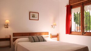 A bedroom with a wooden headboard, a single bed with a striped pillow, two bedside lamps, and a window with red curtains. - 2 -Spectacular apartment with sea view, large terrace, 4 min. walk to the beach (Roses)