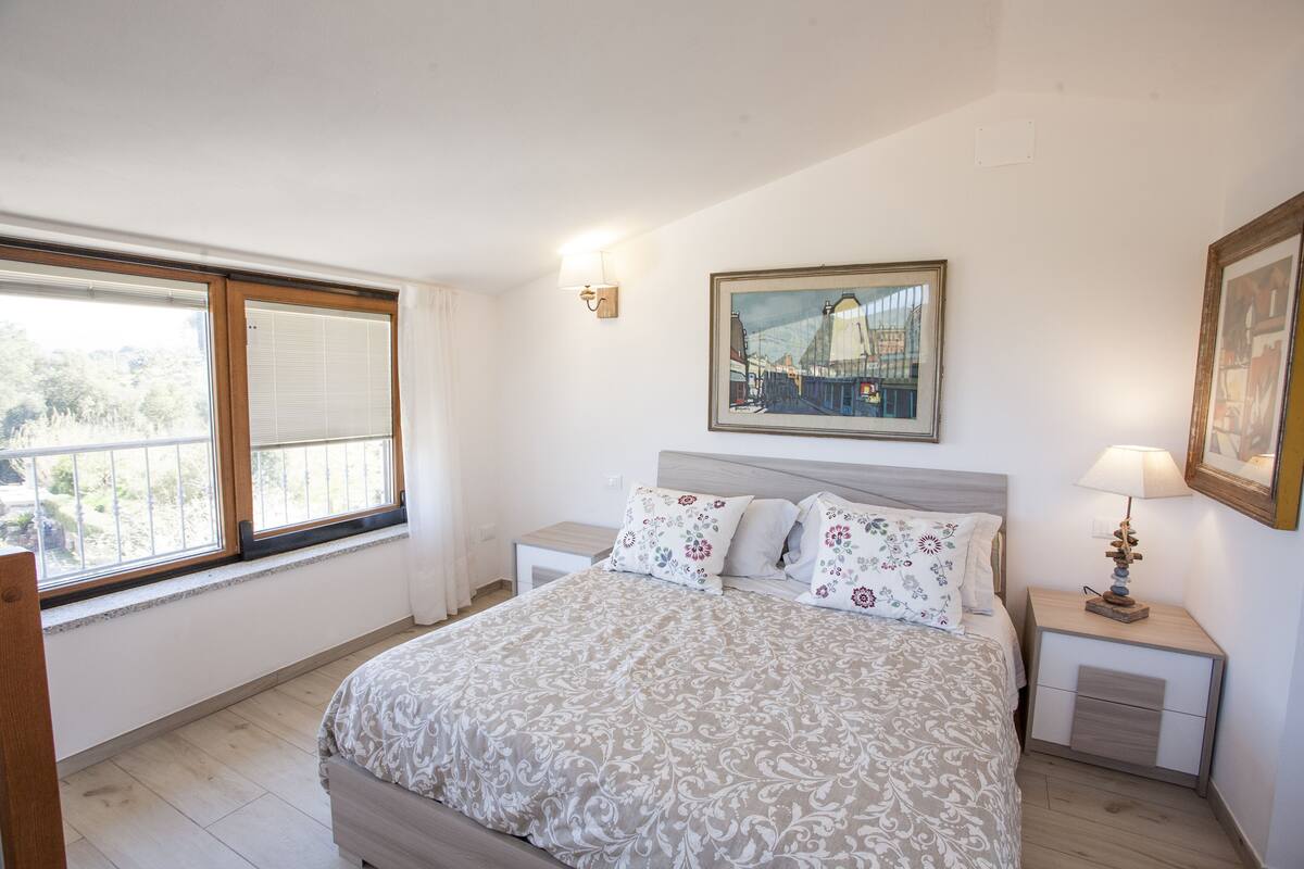 Una camera da letto con un letto, un comodino, una lampada, una finestra con tende e un quadro appeso al muro.