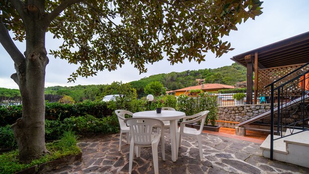 Une terrasse en pierre avec une table ronde et des chaises blanches, entourée de verdure et offrant une vue sur une colline.
