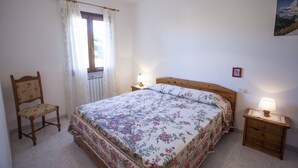 A bedroom with a bed, a chair, a nightstand with a lamp, a window with curtains, and a wall-mounted picture. - Appartamenti Rosa (Campo nell'Elba)