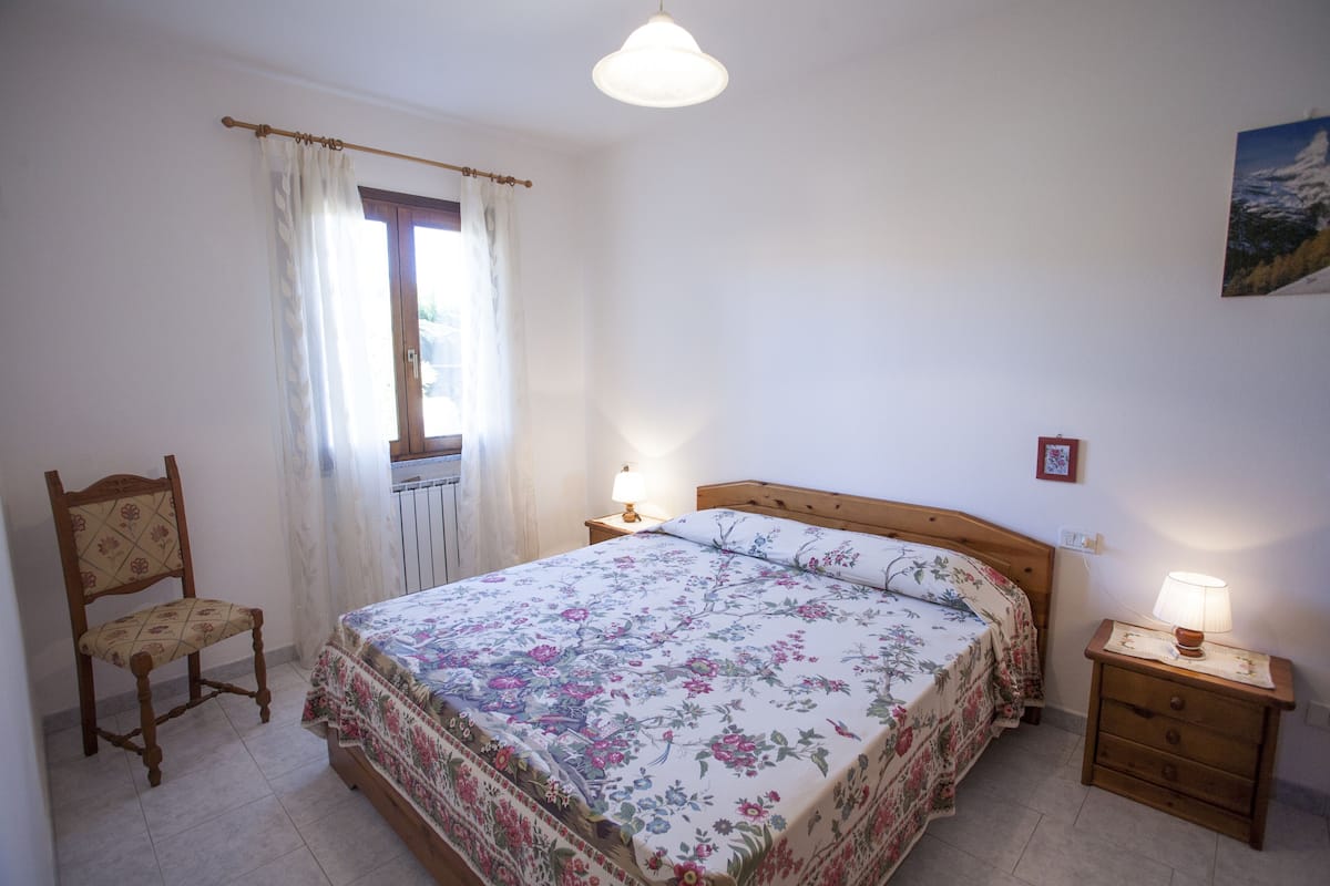 A bedroom with a bed, a chair, a nightstand with a lamp, a window with curtains, and a wall-mounted picture.