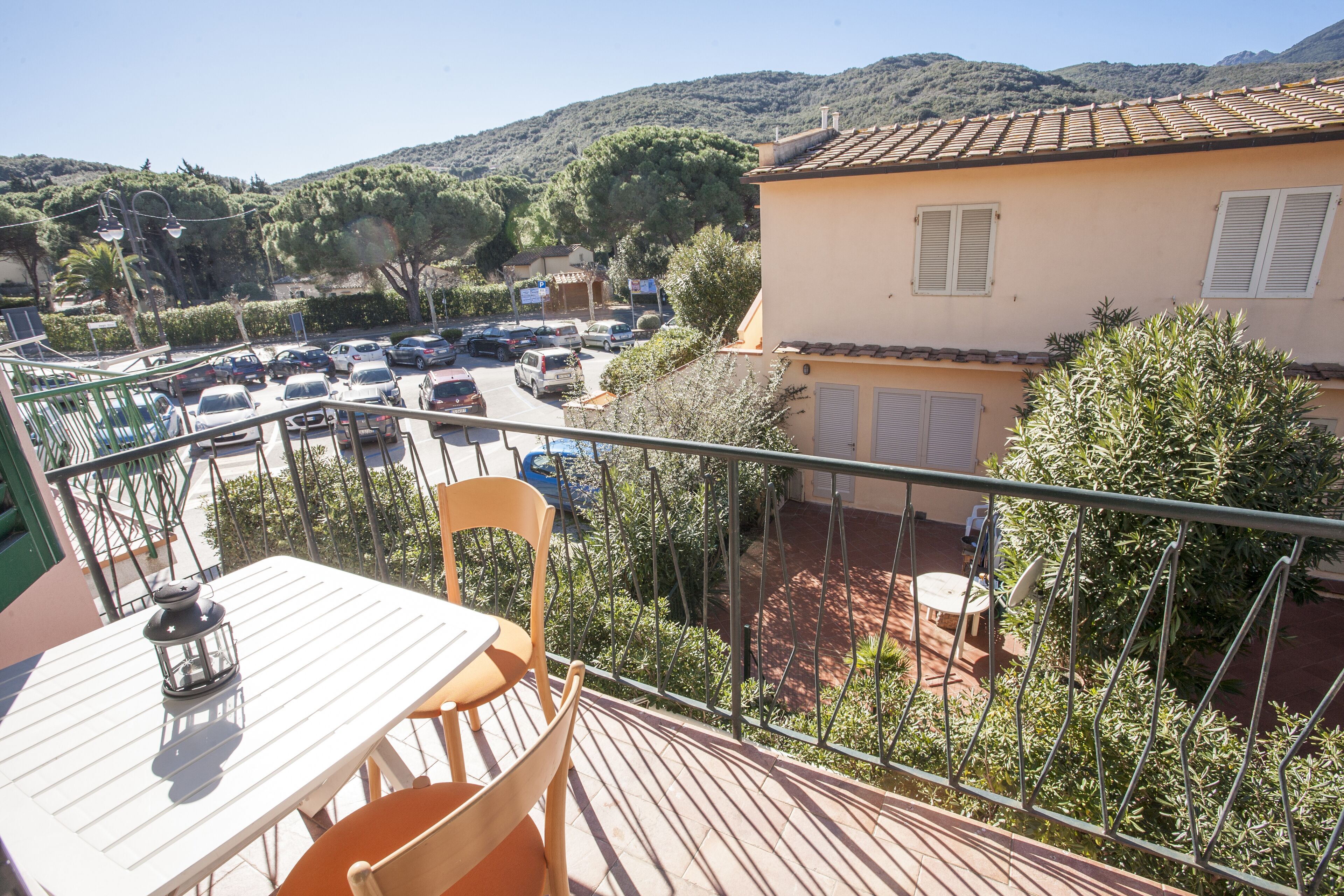 A balcony with a table and chairs, overlooking a parking area and a building with a tiled roof.