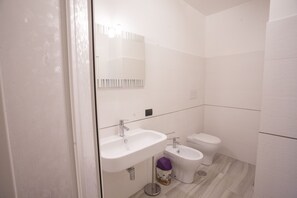 Une salle de bain moderne avec un lavabo, des toilettes et un bidet blancs, agrémentée d’un miroir et d’une poubelle.