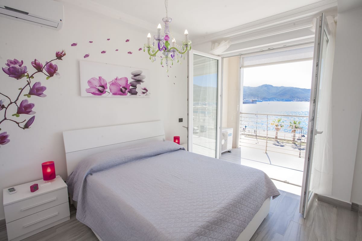 Un dormitorio con cama, mesita de noche, lámpara roja, candelabro y vista a un cuerpo de agua desde un balcón.