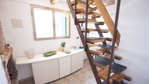 A kitchen with a wooden staircase, white cabinets, a window with blinds, and a tiled floor. - Bilocale Paradiso (Portoferraio)