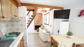 A kitchen with a stove, sink, and wooden cabinets. There is a staircase leading to a loft area with a fireplace. A table with a checkered tablecloth and a potted plant is visible. - Bilocale Paradiso (Portoferraio)