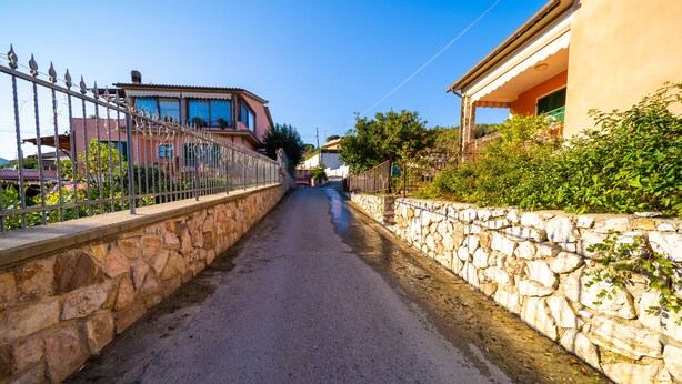 A narrow street flanked by stone walls and metal fences, leading to buildings with large windows and balconies. - Monolocale Nanni (Portoferraio)