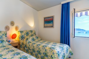 A bedroom with floral bedding, a nightstand with a lamp, a painting, and a window with a view of boats.