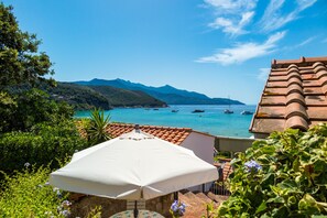 A coastal view with a white umbrella, terracotta roof, and boats on the water.