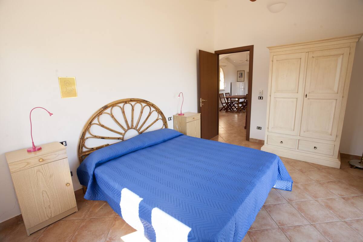 Um quarto com uma cama, uma mesinha de cabeceira de madeira, um guarda-roupa e uma porta que dá para outro cômodo.