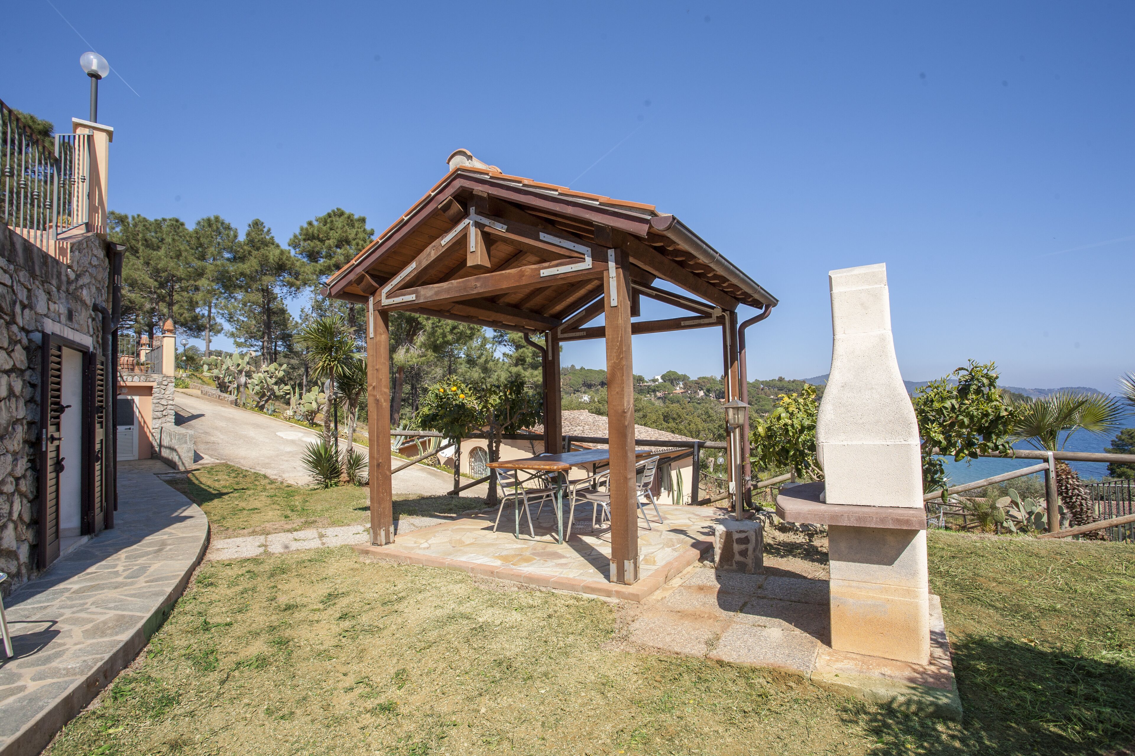 A wooden pergola with tables and chairs, stone steps, and a stone monument in a landscaped garden with trees and a view of the sea.