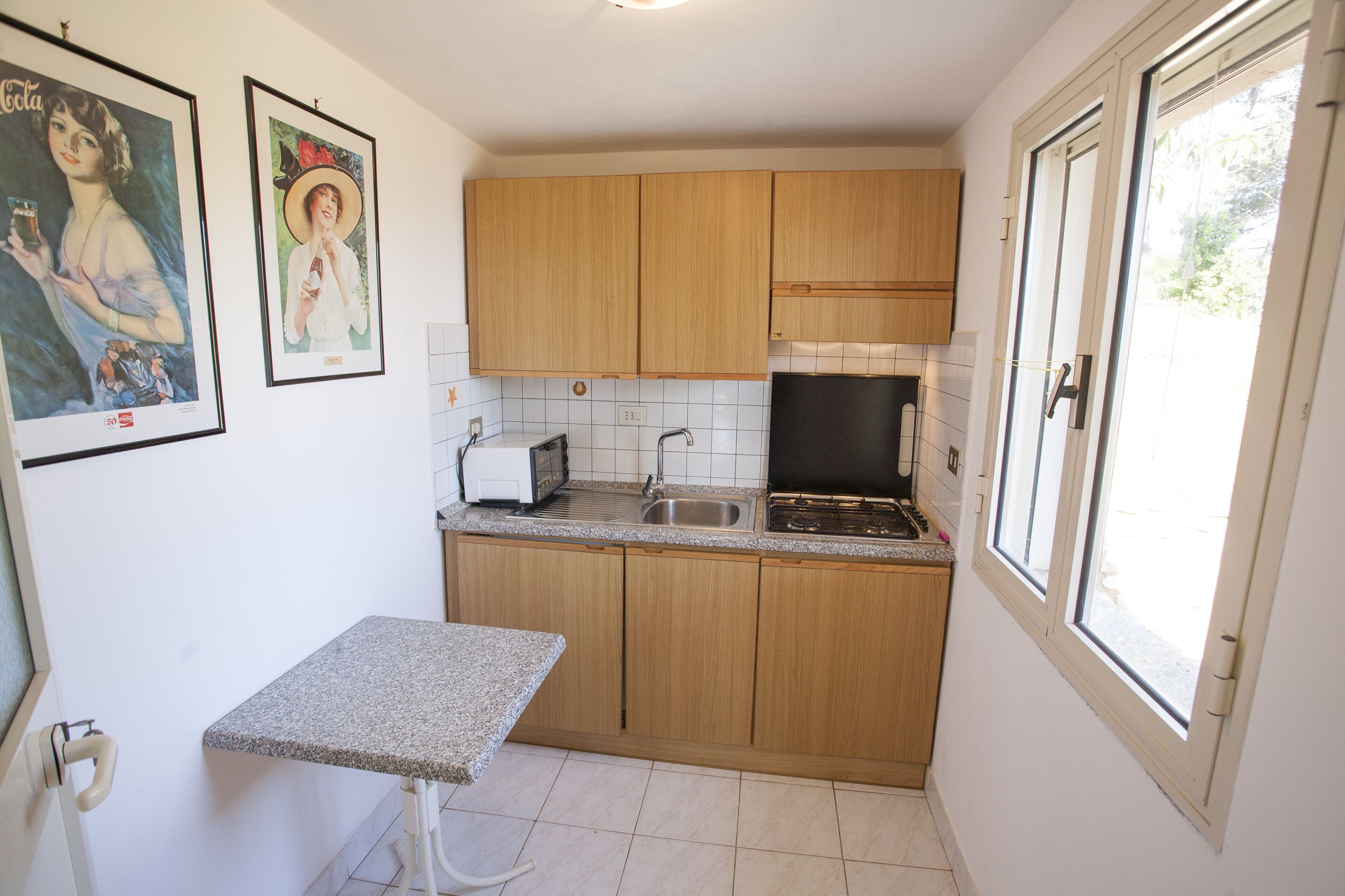 A small kitchen with wooden cabinets, a countertop, a sink, and a stove. There are framed pictures on the wall.