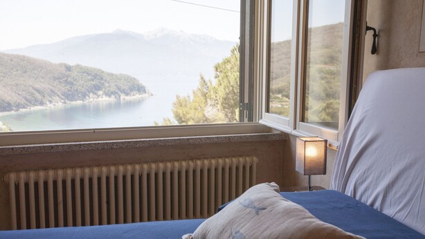A bedroom with a bed, a window with a view of a lake and mountains, and a radiator. - Villetta Paradiso (Portoferraio)