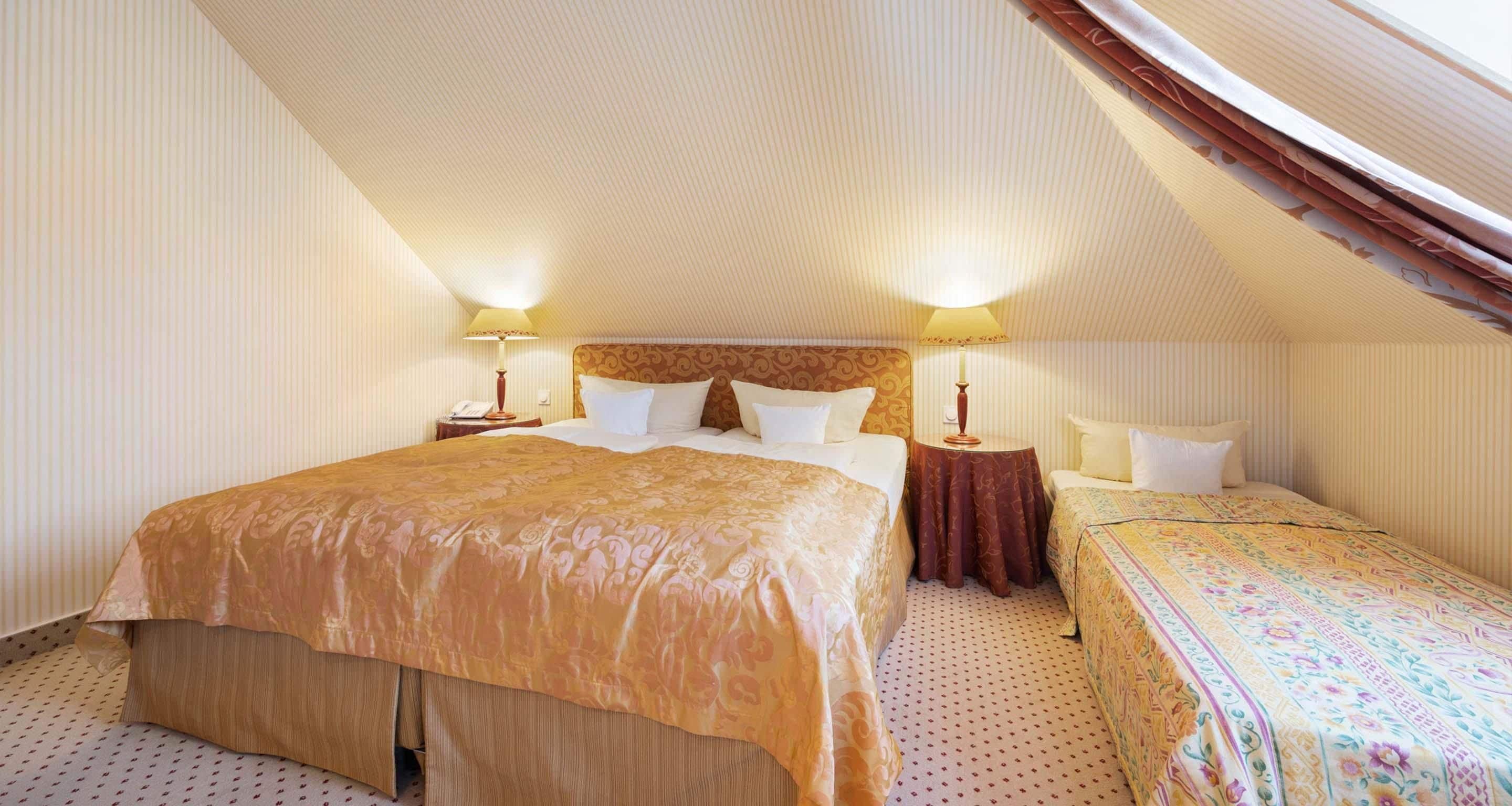 A hotel room with a large bed, a smaller bed, two bedside tables with lamps, and a sloped ceiling.