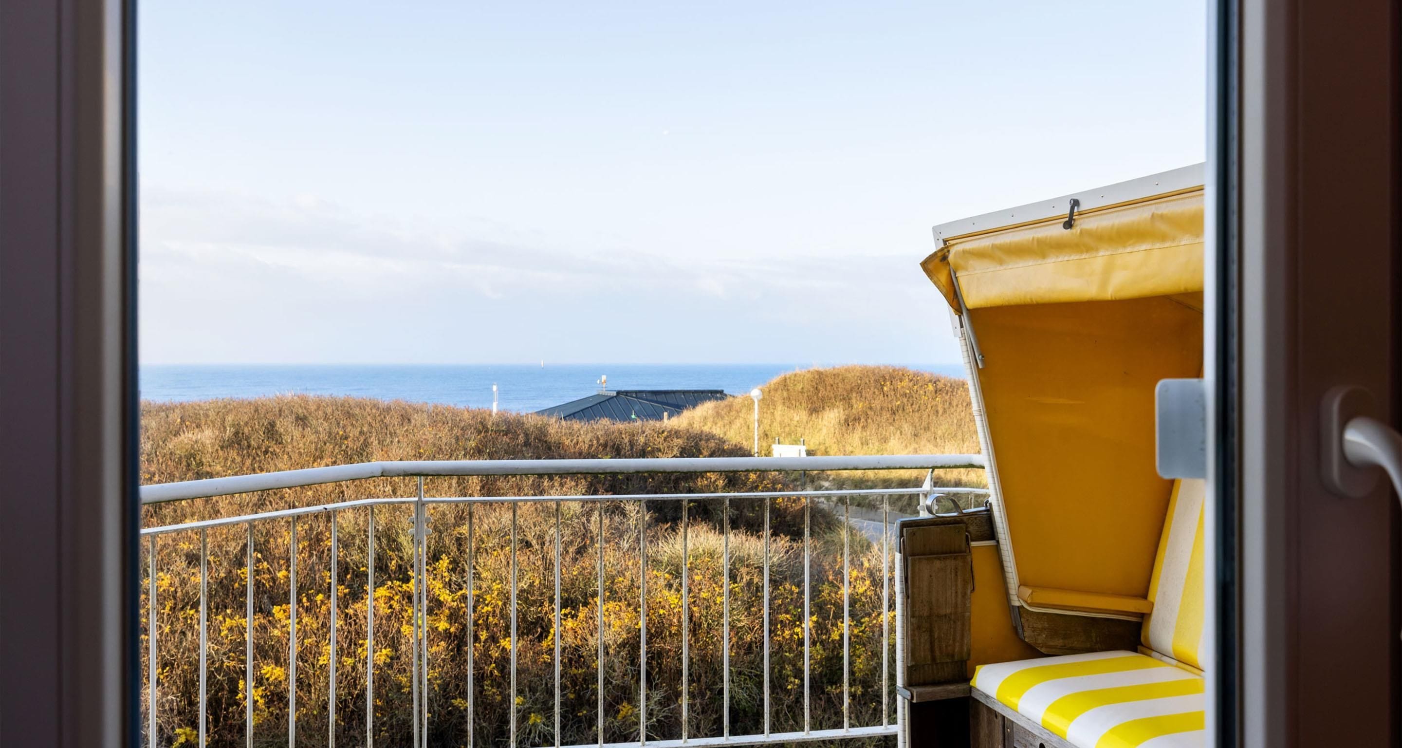 A balcony with a yellow chair, a metal railing, and a view of the sea and hills.