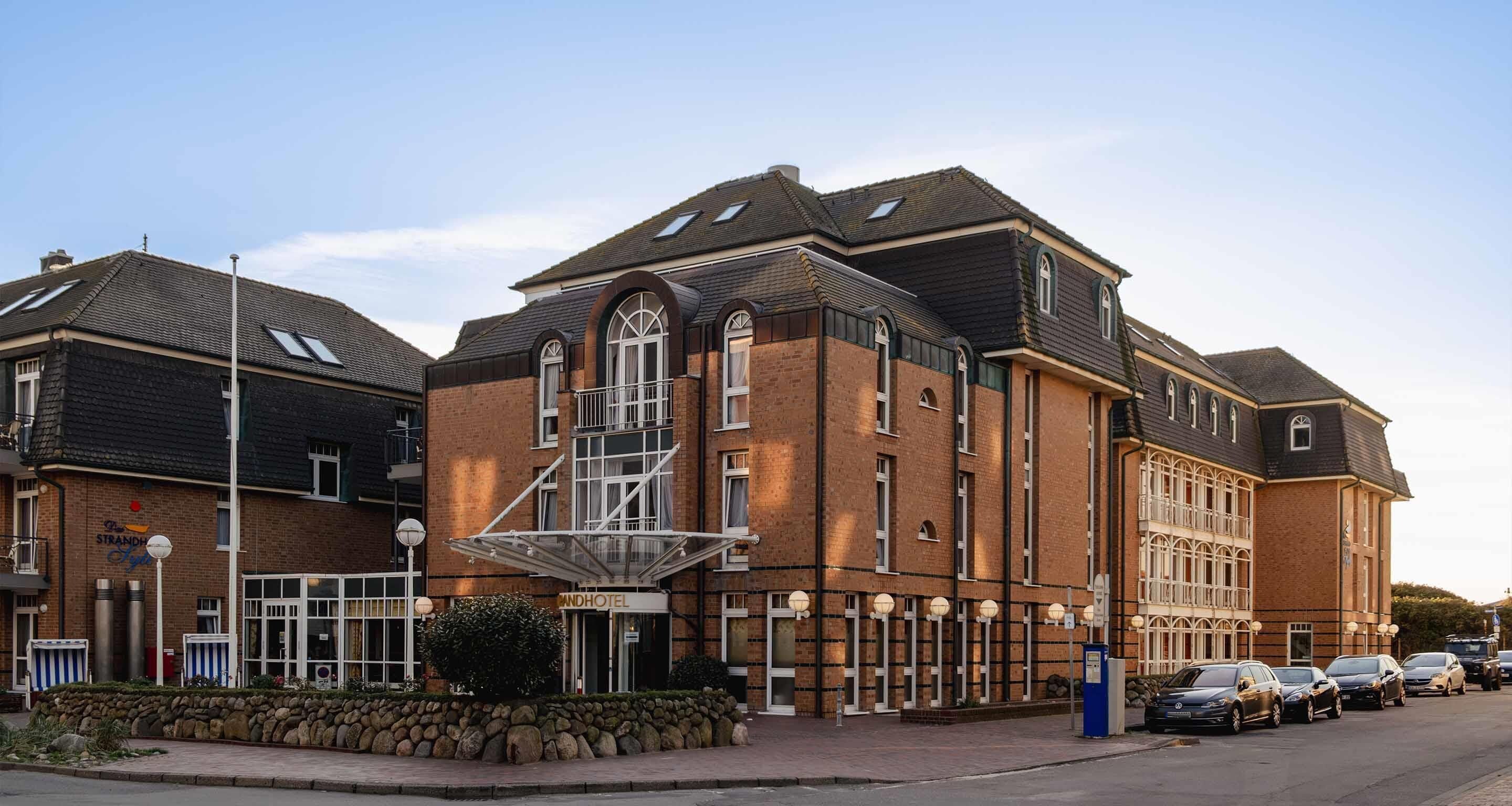 Et hotel med en markant indgang, en trappe op til etageplan og en parkeringsplads med biler.