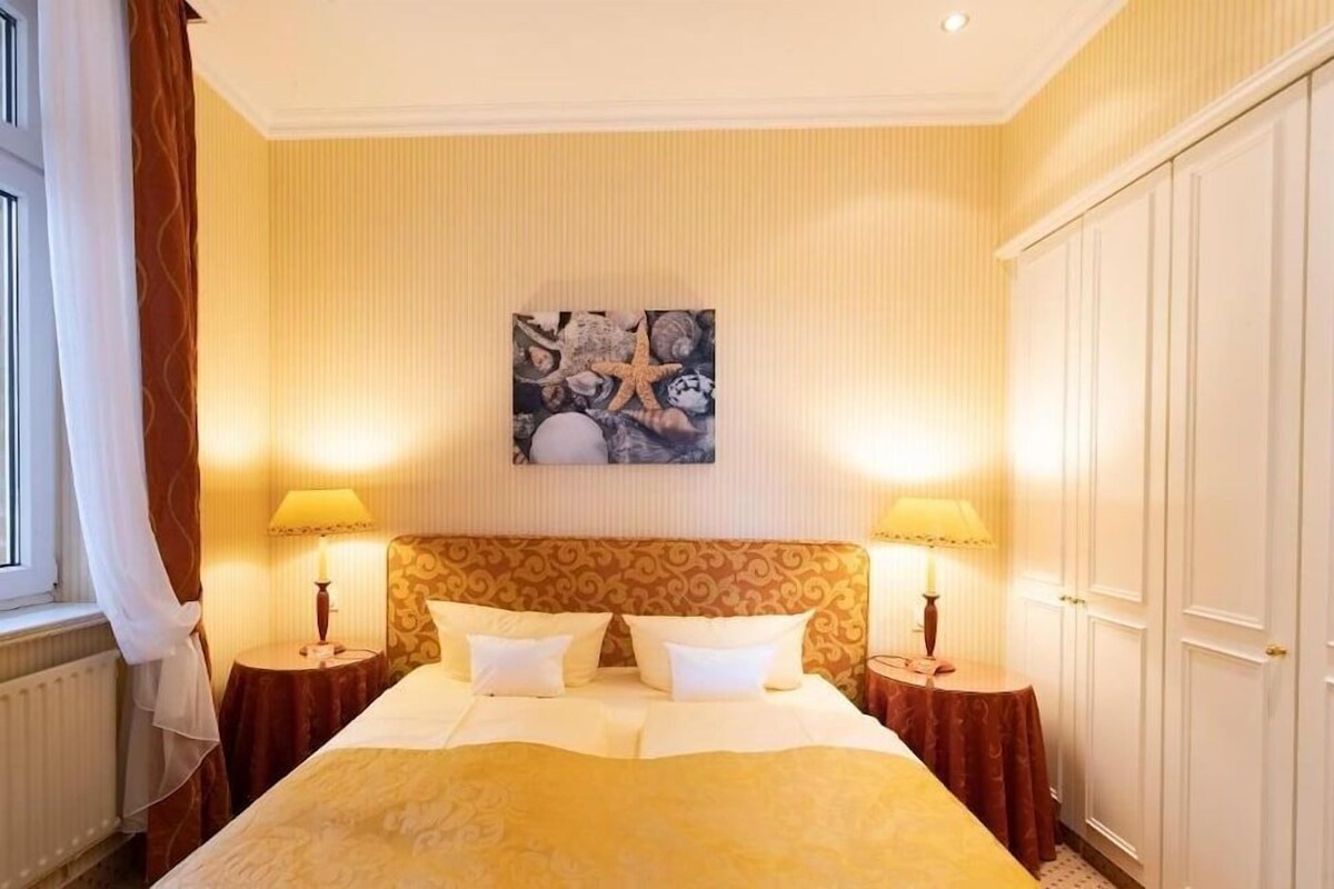 A hotel room with a large bed, two bedside tables, a headboard with a decorative pattern, a wall-mounted artwork, and a wardrobe.