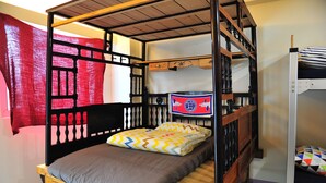 Een stapelbed met houten frame, een enkel bed met een grijs matras en kleurrijke kussens, en een rood gordijn.