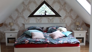 A bedroom with a sloped ceiling, a bed with floral bedding, two nightstands, and a window with a view of trees. - Vacation on the pony farm (Priborn)
