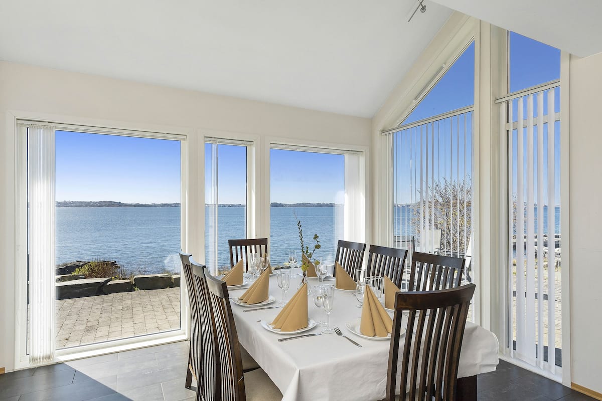 A dining area with a table set for six, large windows with a view of the sea, and a balcony with a railing.