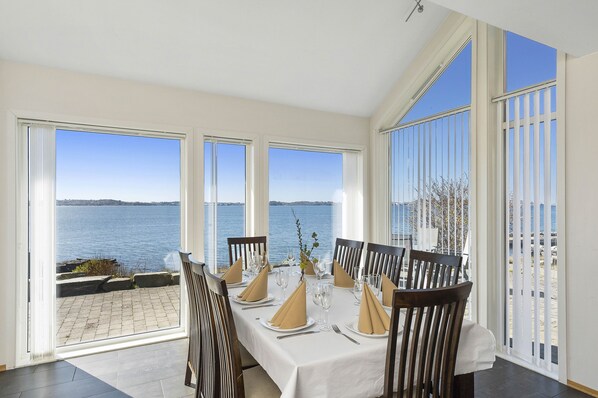 A dining area with a table set for six, large windows with a view of the sea, and a balcony with a railing.