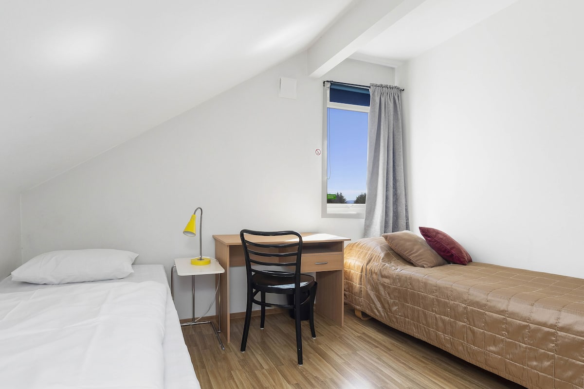 A small, well-lit room with a bed, a desk, and a window with curtains.