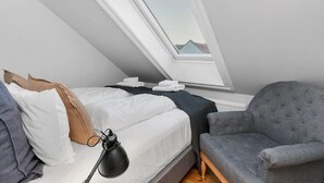Een slaapkamer met dakraam, een bed met kussens, een nachtkastje met lamp en een grijze fauteuil.
